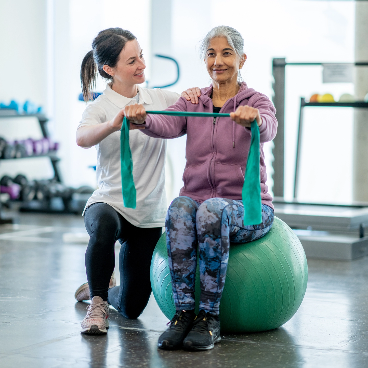 Physiotherapie Balance - Physiotherapie - Eine ältere Frau sitzt auf einem grünen Gymnastikball und zieht an einem Widerstandsband, während eine Therapeutin ihre Schultern führt.