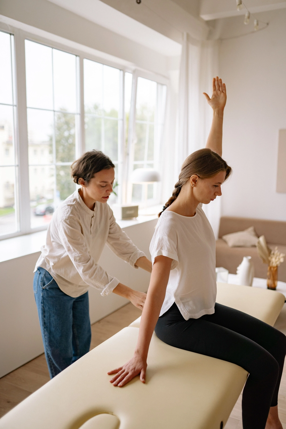 Physiotherapie Balance - Physiotherapie - In einem hellen Studio leitet eine Physiotherapeutin eine sitzende Frau durch eine Dehnübung zur Mobilisierung der Schulter.