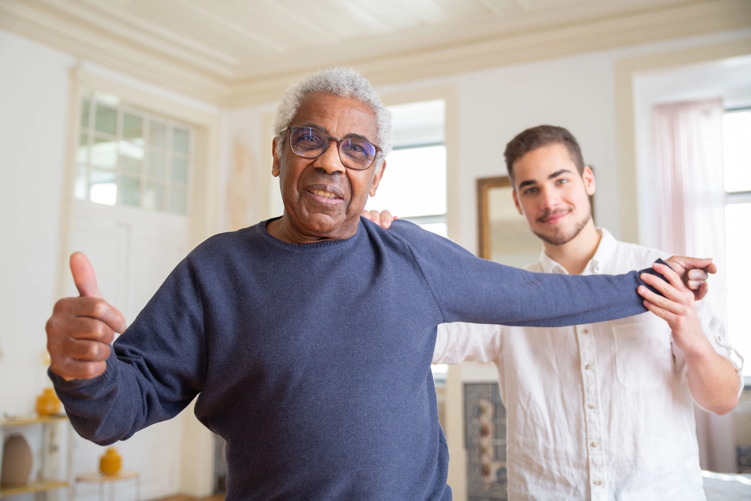 Physiotherapie Balance - Physiotherapie - Ein älterer schwarzer Mann mit grauem Haar und Brille lächelt und zeigt den Daumen nach oben, während ihm ein jüngerer Mann bei Armübungen hilft.
