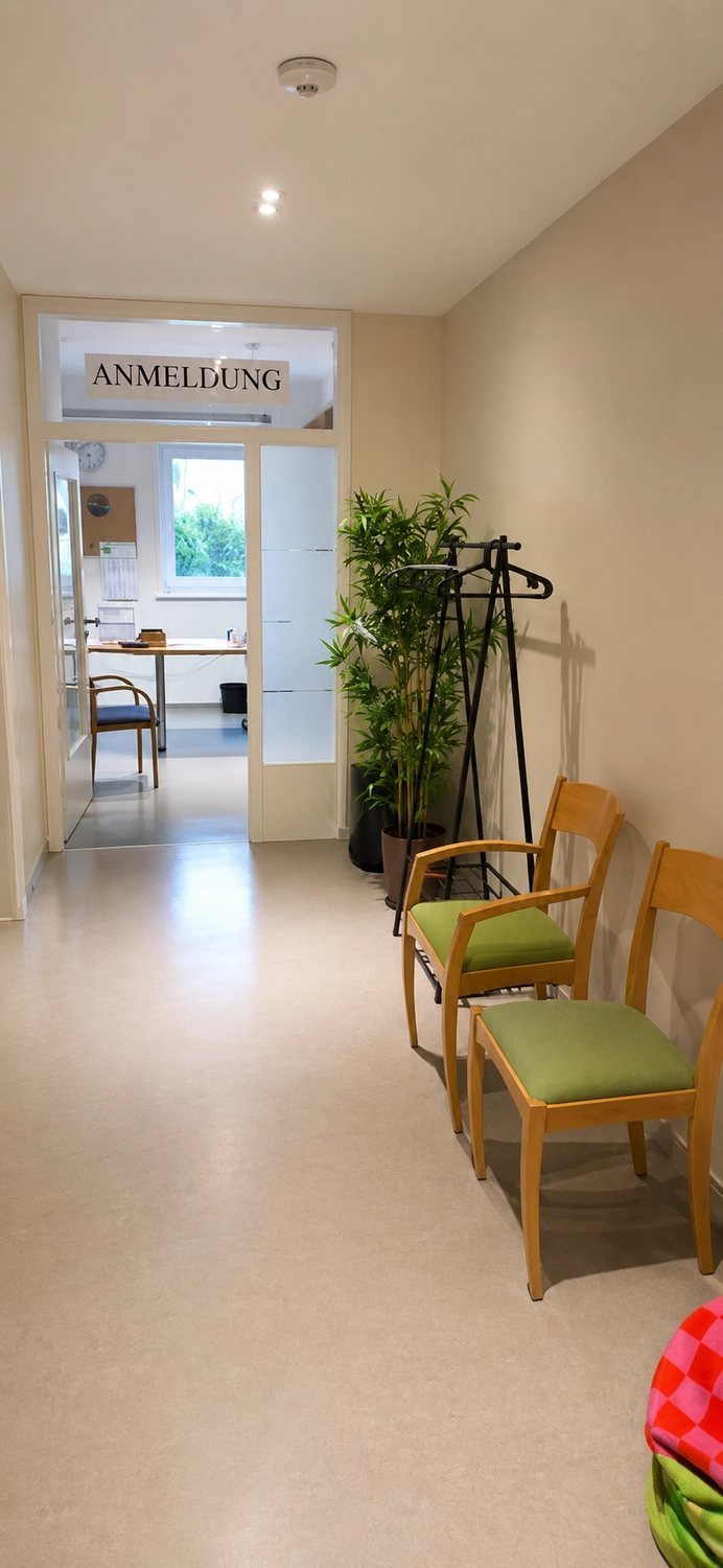 Physiotherapie Balance - Physiotherapie - Ein schmaler, gut beleuchteter Flur führt zu einer Bürotür mit einem Schild darüber, auf dem „ANMELDUNG“ steht. Zwei Holzstühle mit grünen Kissen stehen an der rechten Wand, neben einer schwarzen Garderobe und einer hohen Topfpflanze.