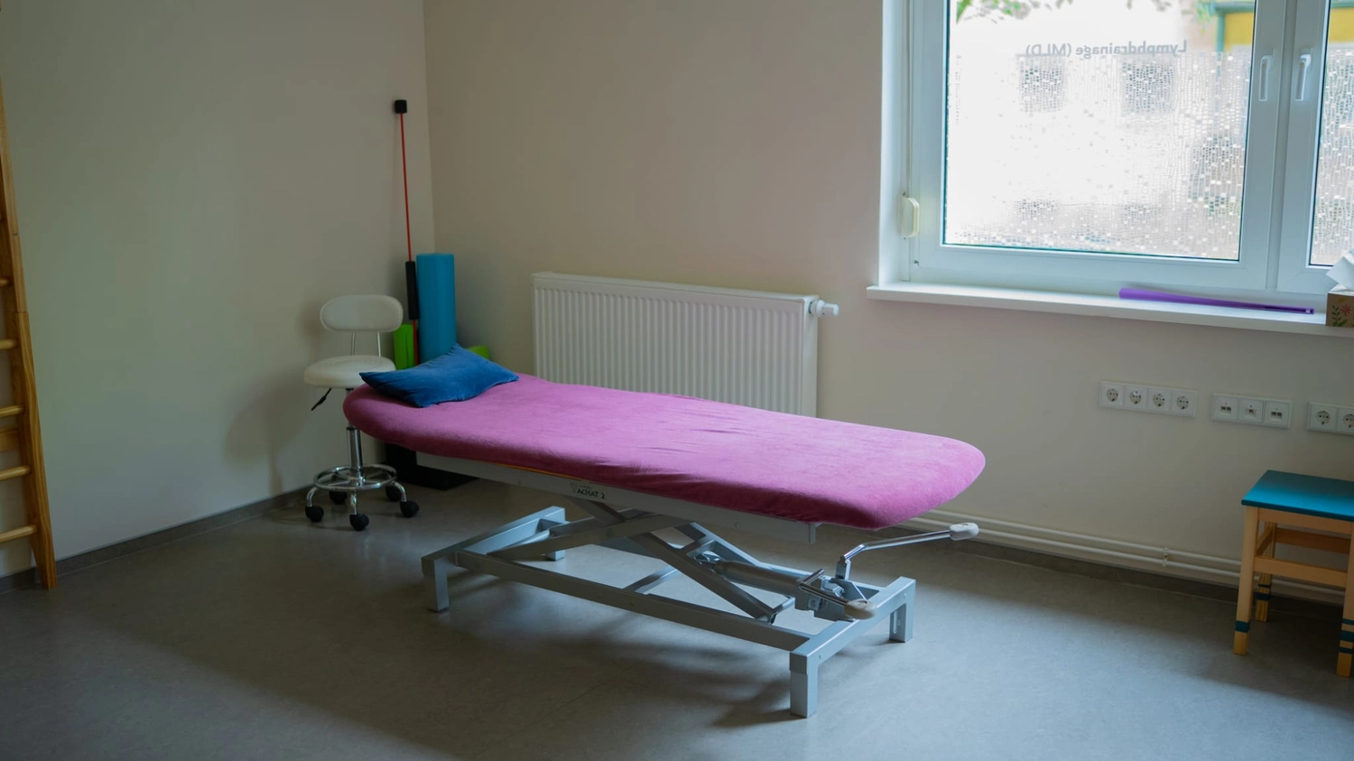 Physiotherapie Balance - Physiotherapie - Auf einem Holzregal neben einer kleinen Lampe und einem Diffusor steht ein rundes braunes Kissen mit gelbem Lotusmuster, auf dem Regal darunter liegt ein rosa gemustertes Kissen.