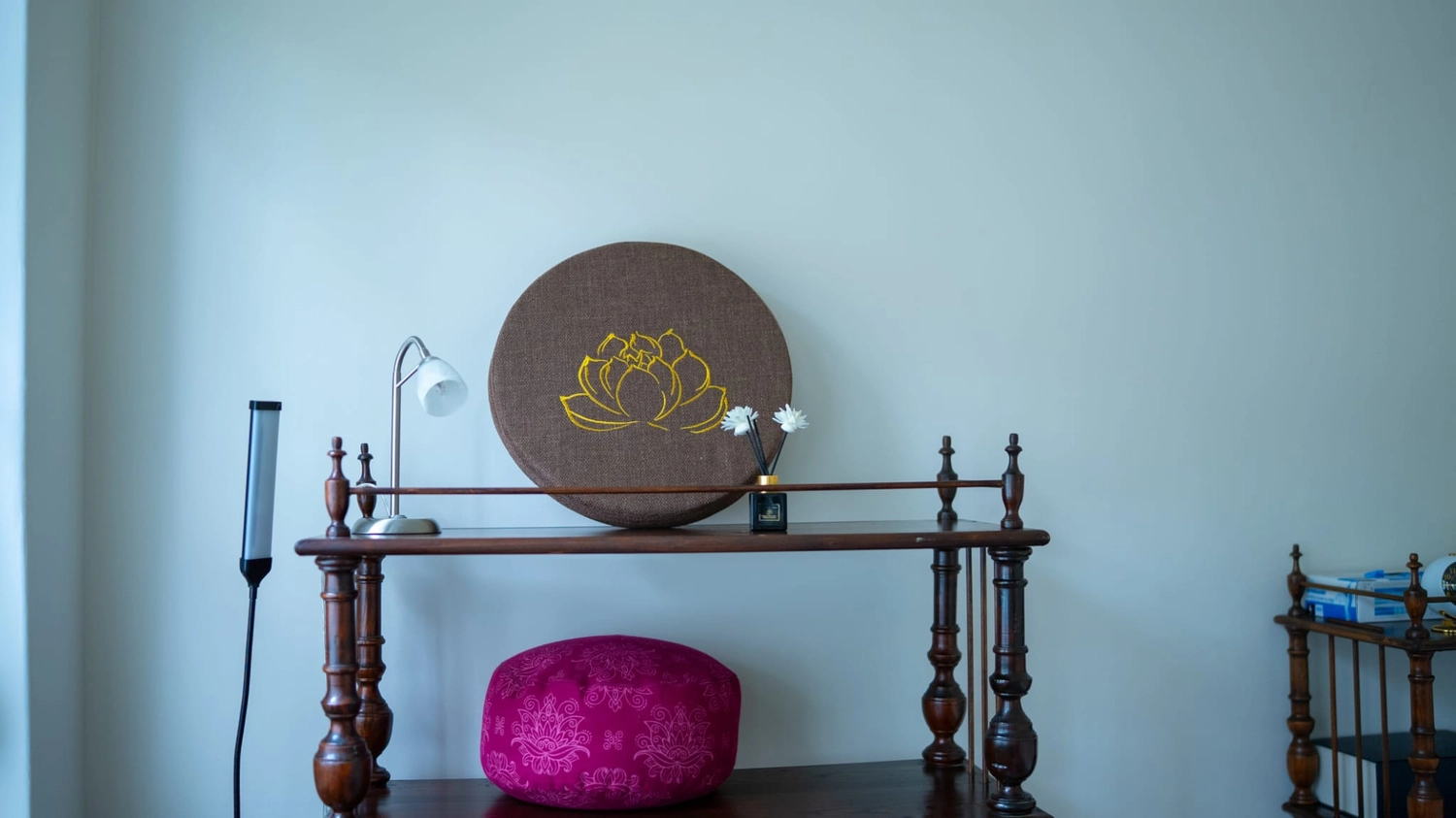  Physiotherapie Balance - Physiotherapie - Ein hölzernes Ausstellungsregal mit einem großen runden braunen Kissen mit gelber Lotusblumenstickerei, einem kleineren rosa gemusterten Meditationskissen und einer Schreibtischlampe.